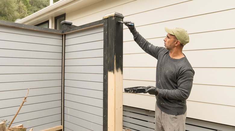 Man wearing long sleeves and a hat painting a wood fence a dark gray color