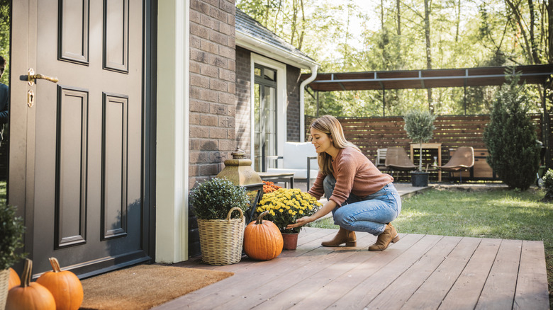 A young woman arranges a porch display with pumpkins and potted fall-flowering plants.