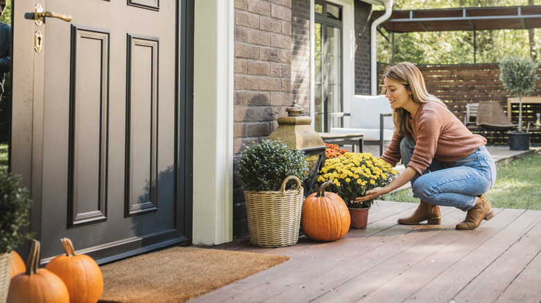 Woman repotting yellow chrysanthemums on a wooden porch near pumpkins and a black door.