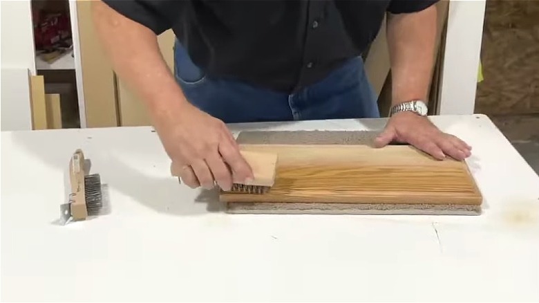 Man's hands brushing an oak cabinet panel with a wire brush to enhance the grain.