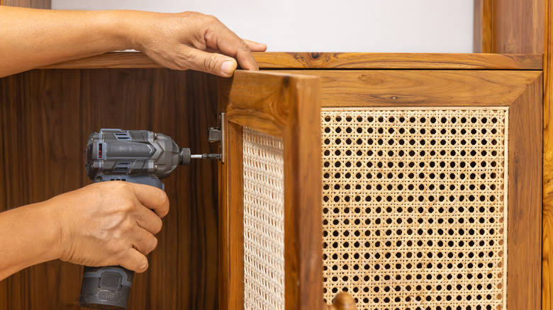 A pair of light skinned hands rehang a caned cabinet door.