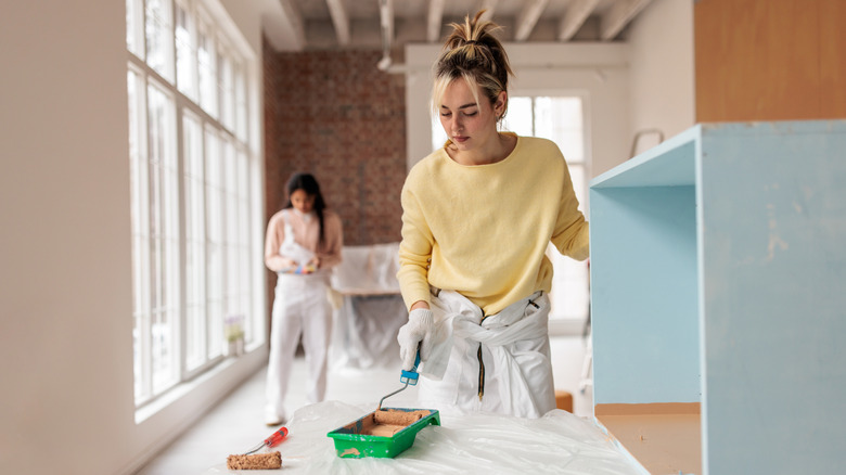 Woman wearing cloves and protective wear, painting cabinet in living room.