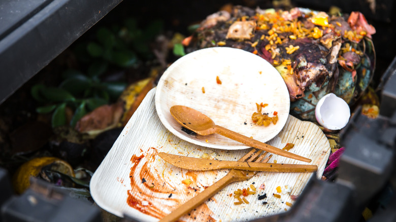 Palm leaf plates and a wooden fork, knife, spoon inside a compost bin filled with food scraps.