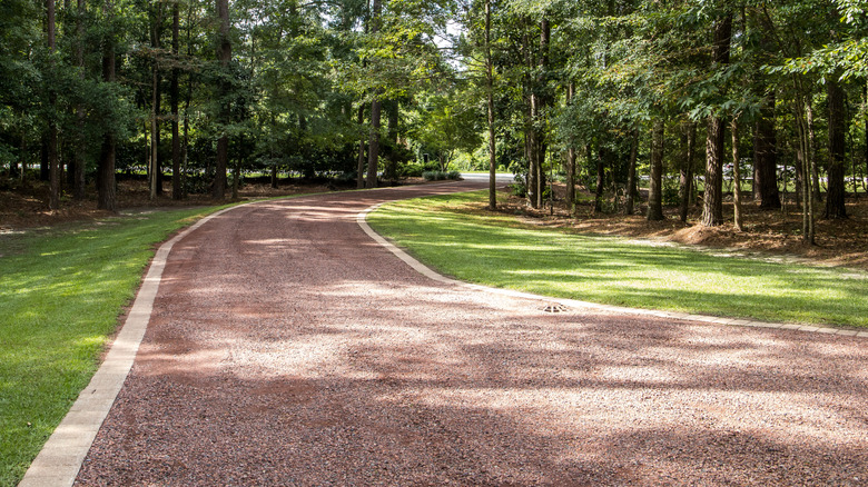 A reddish gravel driveway with a white stone edge running between trees and a manicured lawn