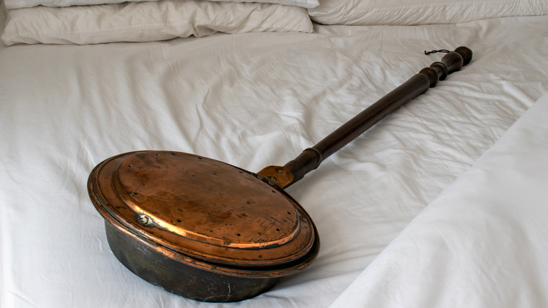 An antique copper bed warming pan with a wooden handle displayed on a bed with white sheets.
