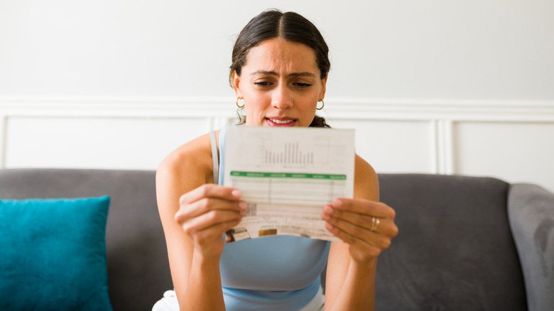 A young woman sits on a sofa holding an energy bill and looking worried.
