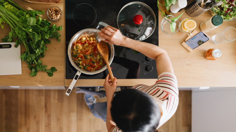 A woman holding a wooden spoon and adding seasoning to a pan of food on an electric stovetop.