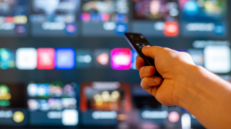 A hand holding a remote, pointing it at a TV with blurry apps in the background
