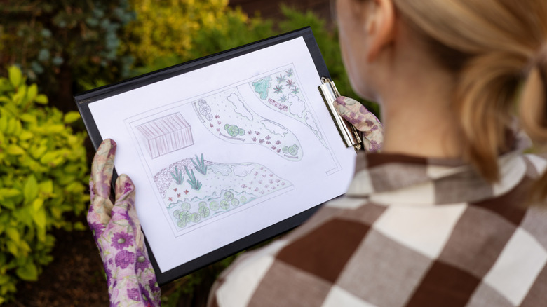 A person wearing gardening gloves looks at a hand-drawn plan for a garden on a clipboard.