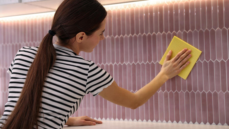 A woman wipes a porcelain tile kitchen backsplash with a dry yellow microfiber cloth.