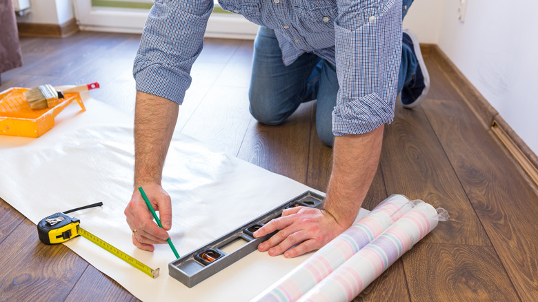 A man measures wallpaper for cutting on a wood floor.