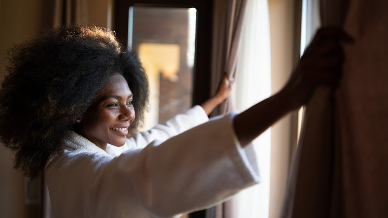 Smiling person in a bathrobe opens curtains