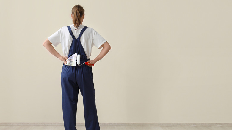 A woman wearing a white t-shirt and blue overalls, holding paint swatches and a paint roller, has her back to the camera and is staring at a tan-colored wall.