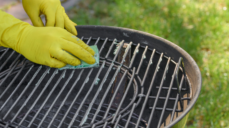A yellow-gloved hand cleaning a grill with a sponge