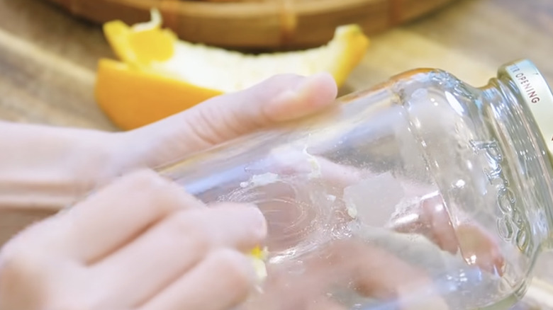 A person uses lemon peels to remove the sticker residue from a glass jar.