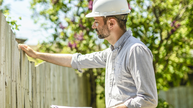 A man with a hardhat and a clipboard inspects an old wooden fence that needs to be removed.