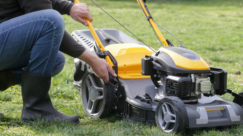 Person kneeling next to a yellow and black push lawn mower and adjusting its height