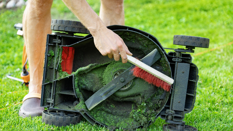 A man brushing clumped grass off of lawnmower blades.