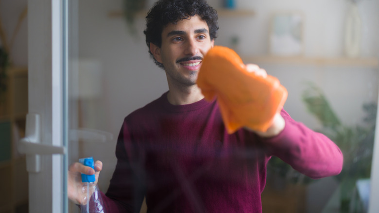 A young homemaker smiles while washing a window with an orange rag