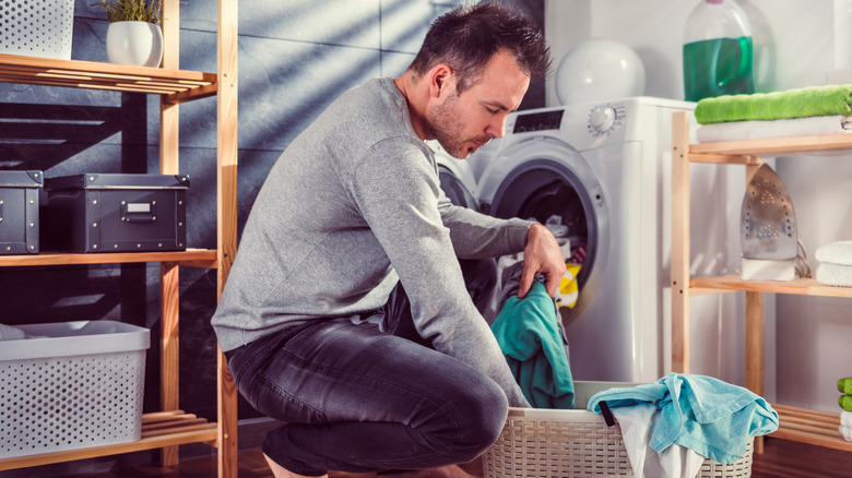 A man wearing a gray sweater takes clothes out of a laundry hamper near a washing machine at home