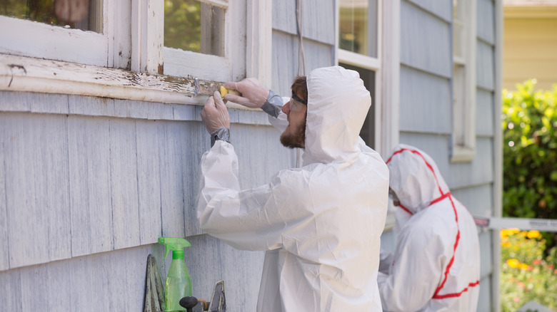 Two people in hazmat suits removing lead paint from the exterior of a pale blue home