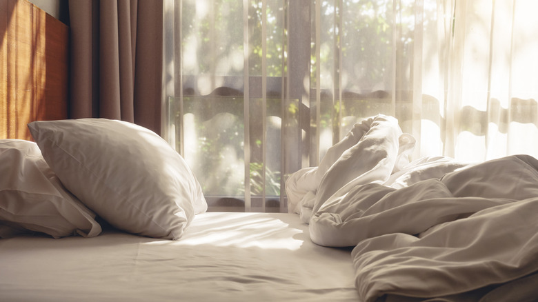 A bed with crumpled white bedsheets and sunlight filtering through a sheer curtain
