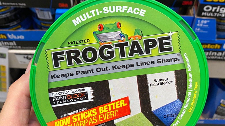A hand holds a roll of of green FrogTape in front of a hardware store shelf.