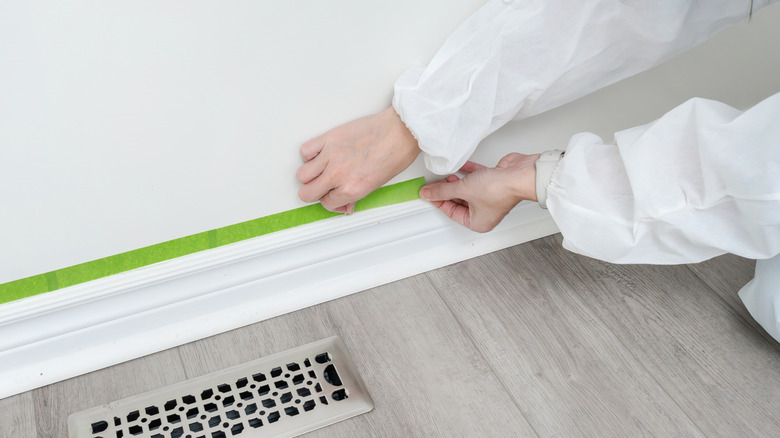 A person applies green painter's tape to the top of a baseboard on a white wall in a home.