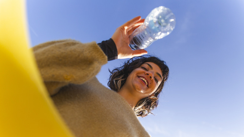 A smiling person viewed from below tosses a plastic bottle into a recycling bin.