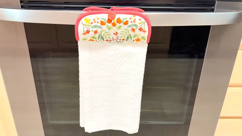 Colorful pot holder sewn to a white dish towel hanging on bar on stainless steel oven