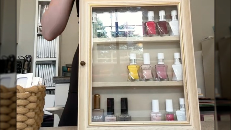 A person holds a shadow box with a glass door and nail polish on the shelves.