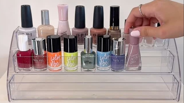 A transparent acrylic shelf riser stand with a hand adding a nail polish bottle to one tier.