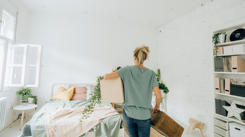 A woman holds storage boxes in her hands as she arranges furniture and decor in a bedroom.