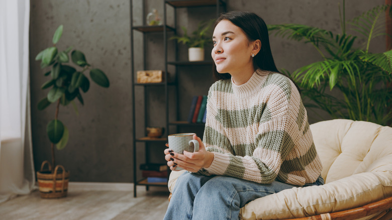 A woman sitting in a parlor cupping a hot drink in her hands.