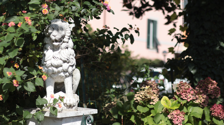 a garden statue of a lion on a pedestal is framed by blooming shrubs
