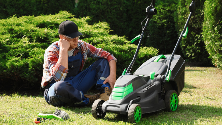 A man sits near a lawnmower that is not working