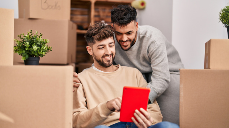 A smiling gay couple sit among moving boxes and look at a tablet together.