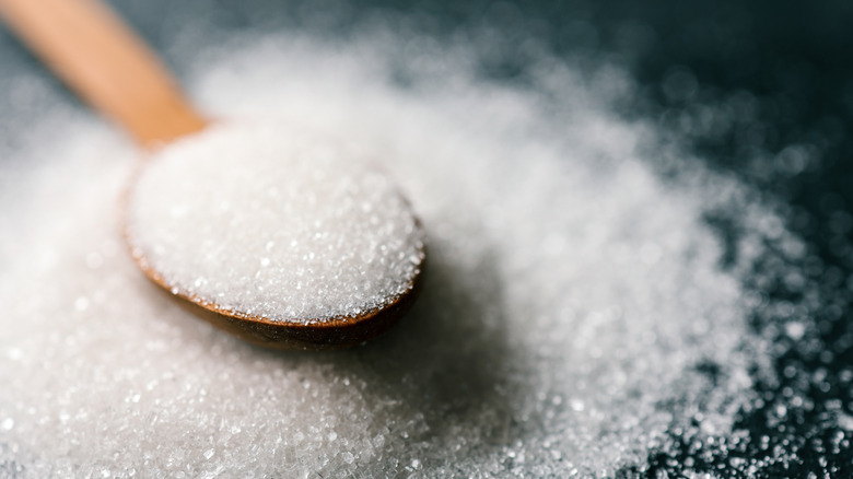 Scoop of granulated white sugar in a wooden spoon, the scoop overflowing onto a black countertop surface
