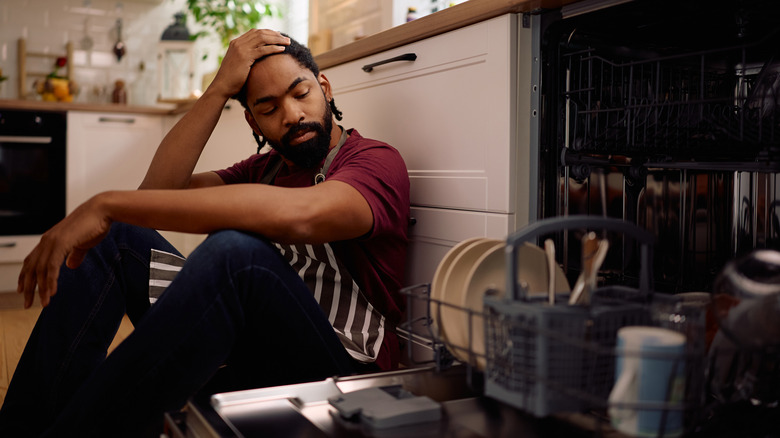 A man holds the top of his head while sitting next to an open dishwasher
