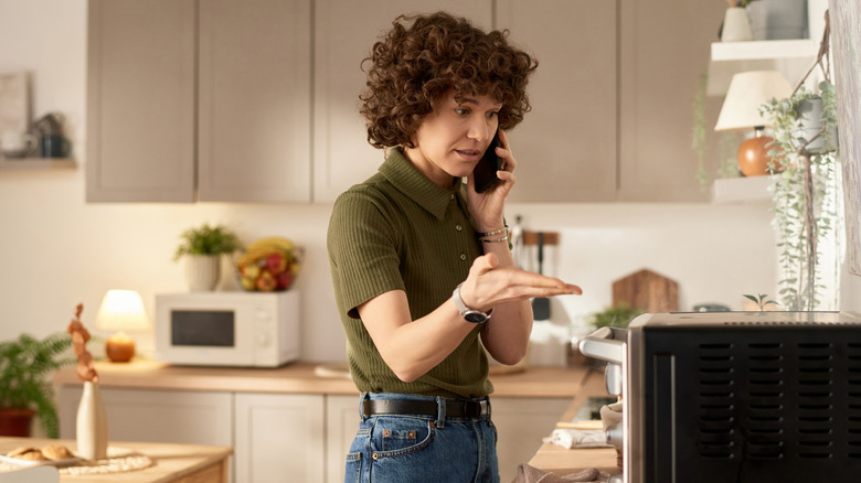 A woman on the phone in the kitchen gestures in front of an appliance
