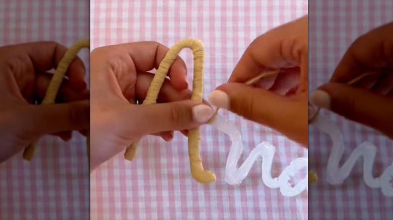 A pair of women's hands wraps raffia ribbon around letters made from hot glue.