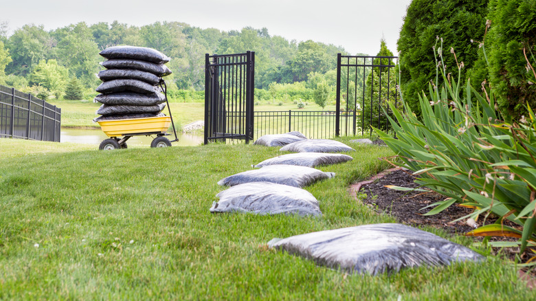 A yellow cart with bags of mulch and bags of mulch laying beside a garden bed waiting to be spread out.