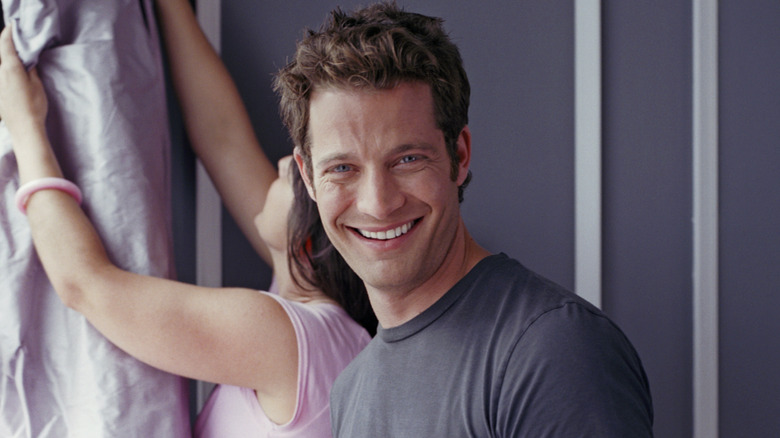 A headshot of Nate Berkus smiling at the camera with another person hanging curtains behind him