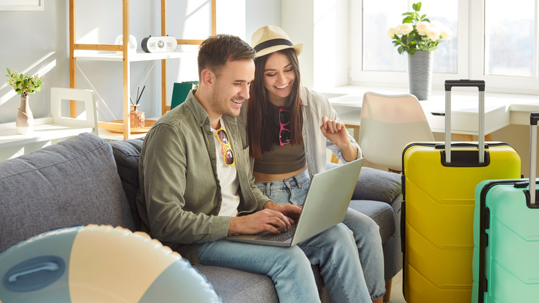 A young couple sitting on a couch in a living room with luggage looking at a computer and smiling