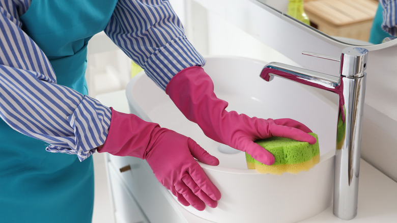 Pink gloved hand cleaning bathroom sink with sponge.