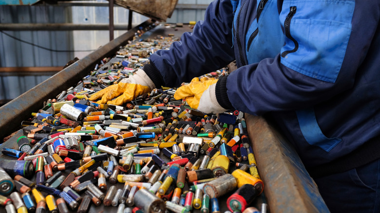 Inside a hazardous waste facility, a worker in a blue jacket and yellow gloves is handling numerous used batteries