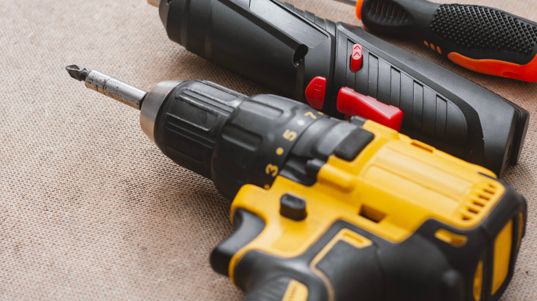 Set power tools including yellow cordless drill with battery pack, electric screwdriver, and manual screwdrivers handles on a wooden background.