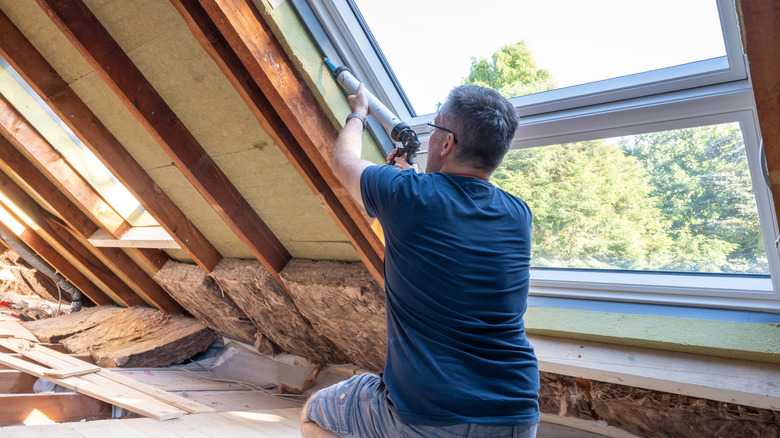 A person applies caulk to the perimeter of a window in an attic