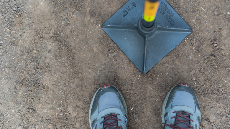 A man wearing sneakers uses a tamper to compact soil.