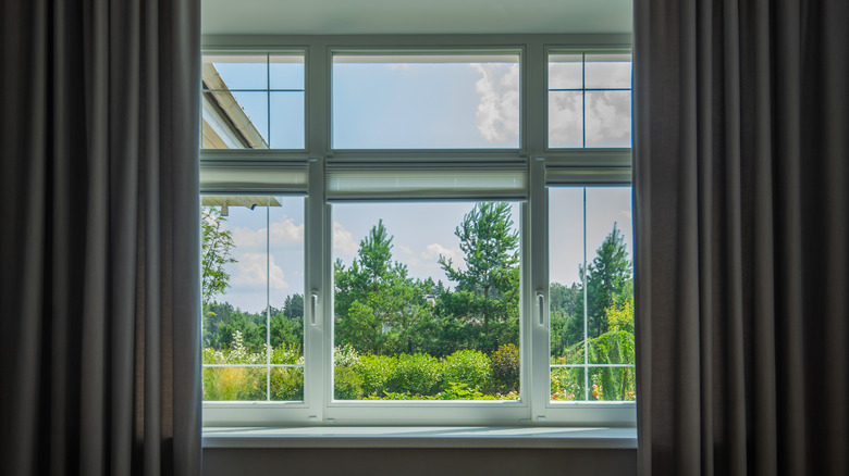 Window with grey drapes looking out on pastoral scene with trees and shrubs.